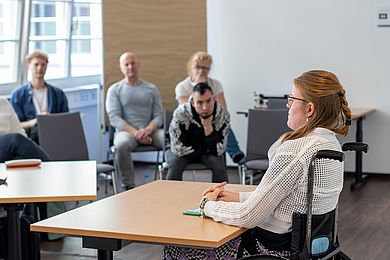 Eine Frau sitzt an einem Tisch im Rollstuhl und spricht vor einer Gruppe von Menschen, die im Hintergrund zu sehen sind und auf Stühlen sitzen.