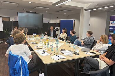 11 Personen sitzen um einen länglichen Tisch herum in einem großen Büroraum. Sie hören einem Mann zu, der gerade spricht. Auf dem Tisch liegen Notizblöcke und Stifte verteilt und Wasserflaschen stehen in der Mitte. 