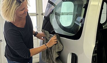 Eine Frau poliert mit einem weichen Tuch die rechte hintere Seite eines Autos. 
