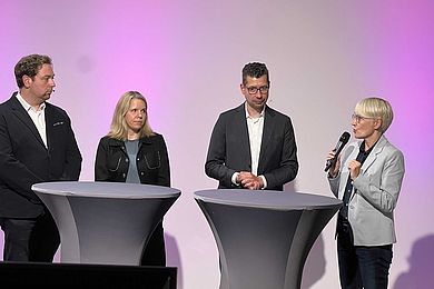 Studioansatz von vier Expertinnen die nebeneinander an 2 Stehtischen platziert sind. Eine Dozentin spricht in das Mikro. 