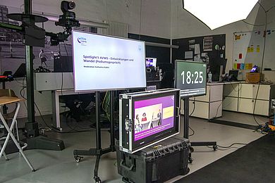 Studioansicht aus Sicht der Referierenden. Sie sehen auf 3 Bildschirme. Mitte oben gibt die Präsentation wieder. rechts gibt die Zeitdauer des Beitrages wieder und unten mittig ist die Live-Streamin-Ansicht der gesamten Veranstaltung zu sehen. Ganz im Hintergrund hängt die Kamera an einem hohen Stativ und es sind mehrere Techniker-Tische zu sehen. 