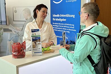 Messestand der EAA Sachsen, eine Frau mit kurzen Haaren, Brille und weißer Bluse hinter einem Counter. Die Frau ist im Gespräch mit einer anderen Dame mit kurzen Haaren und Brille, die eine helltürkisene Jacke trägt.