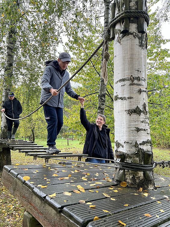 Ein Teilnehmer balanciert auf einem Seil und erhält Unterstützung von einer Frau, die nebenher läuft und ihm die Hand reicht. Das Seil ist zwischen Bäumen gespannt.