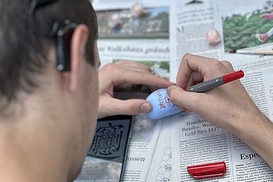 Einer der Teilnehmenden beschreibt einen blau angemalten Stein mit einem roten Filzstift. Hinter seinem Ohr trägt er ein Hörgerät.