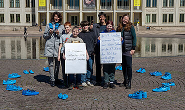 Eine Gruppe erwachsener Personen und Jugendlicher steht in einem großen Kreis aus leuchtend blau angemalten Schuhen. Sie lächeln in die Kamera und halten zwei Plakate in die Höhe.