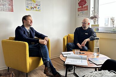Zwei Personen sitzen in einem hellen Raum in gelben Sesseln und tauschen sich in entspannter Atmosphäre aus. Auf dem Tisch stehen Wasser und Unterlagen, an den Wänden hängen Informationsplakate.