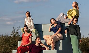 Zu sehen sind sechs junge Frauen auf einer hellgrünen Treppe mitten auf freiem Feld vor blauem Himmel