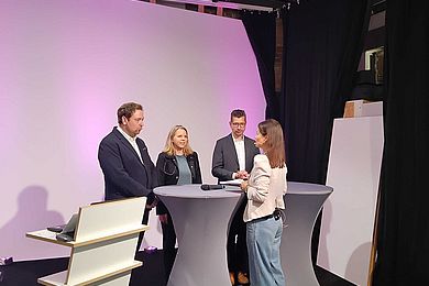 Studioszene mit der Expertinnenrunde. Die Moderatorin gibt den Dozentinnen Informationen. Alle stehen um Stehtische herum. 