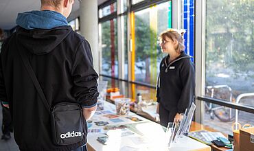 Eine Person ist scharf im Vordergrund von hinten zu sehen. Sie steht an einem Informationstisch mit verschiedenen Flyern und Informationsblättern. Hinter dem Tisch im Foyer des Berufsbildungswerks Leipzig steht eine*r Mitarbeiter*in, unscharf zu sehen.