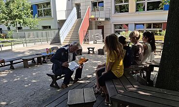 US-Konsul Dave Panetti liest Kindern aus dem Buch "Die kleine Raupe Nimmersatt" vor. Die Kinder sitzen ihm gegenüber auf einer Bank unter einem Baum. 